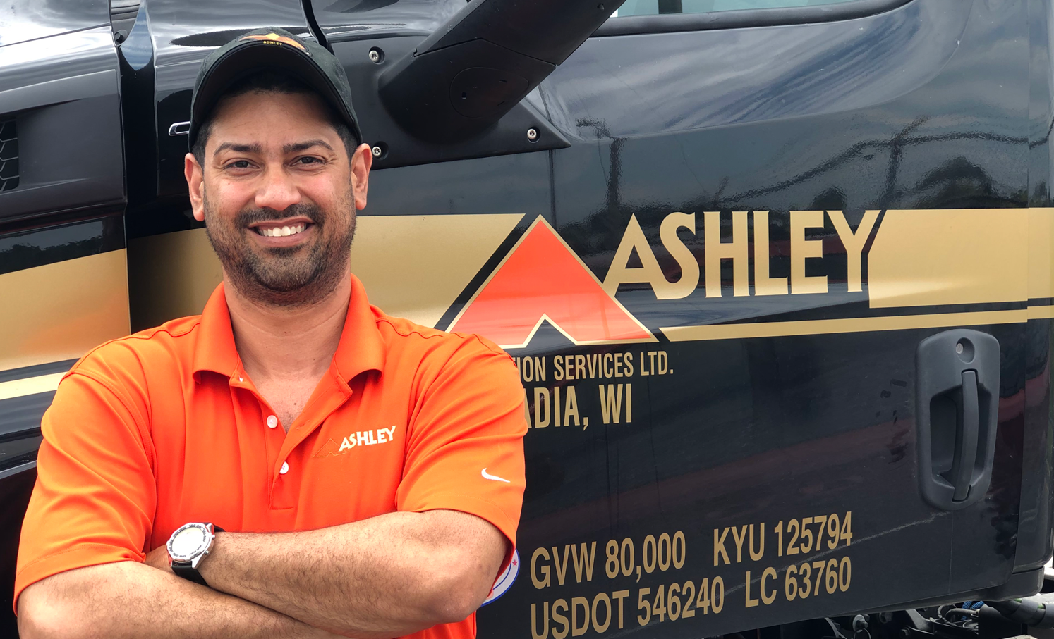 Ashley Driver with an orange shirt, has his arms crossed in front of an Ashley Truck