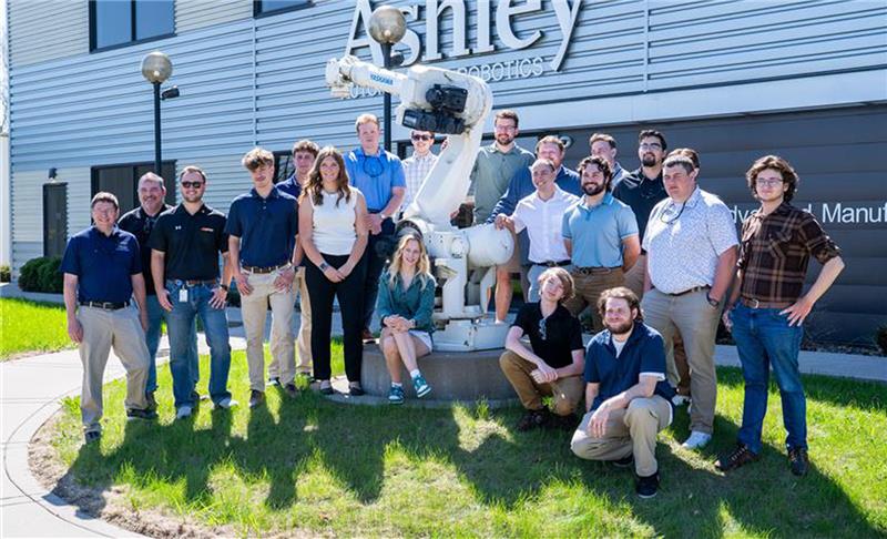 A group of people are next to an Ashley Robot arm smiling at the camera